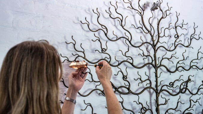 A figure with long hair using a screw driver to attach a metal leaf to a metal tree sculpture